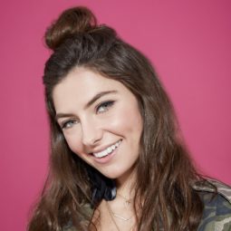 Casual hairstyles: Brunette model with shoulder length wavy hair in a half-up bun style, wearing a green camo jacket and standing in front of a pink background