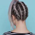 coloured braids: back shot of model with lilac hair styled into upside down French braided ponytail, wearing stripes and posing in a studio setting