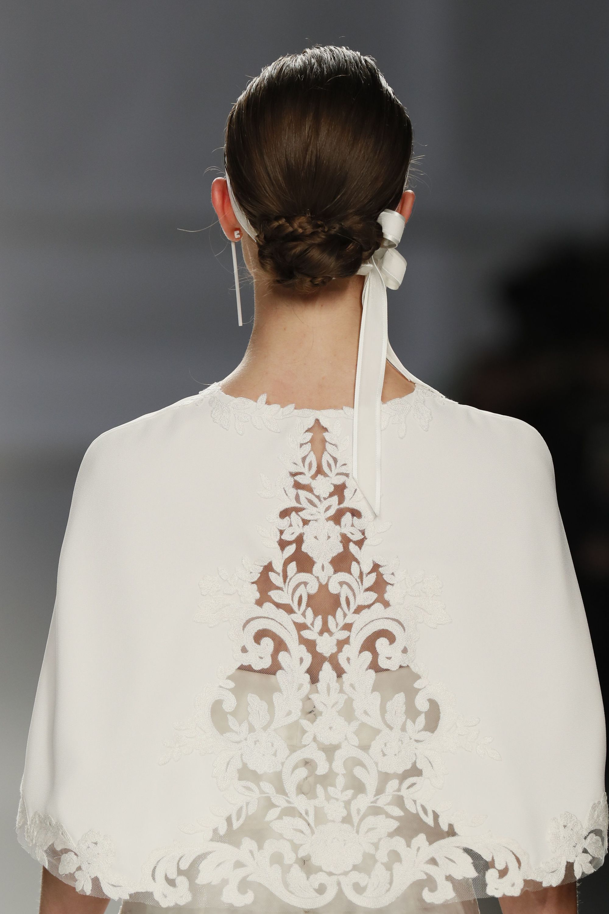 Wedding updos: Close up shot of model on the bridal runway with a braided low bun, complete with white ribbon around, wearing a white wedding cape and drop earrings