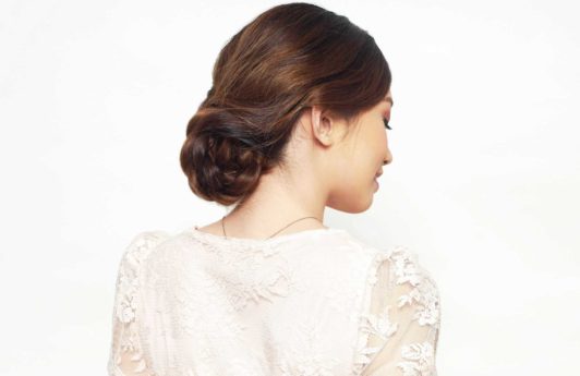 Easy prom hairstyles: Back shot of a model with chestnut brown hair styled into a low braided updo, wearing a white lace dress and posing in a studio.