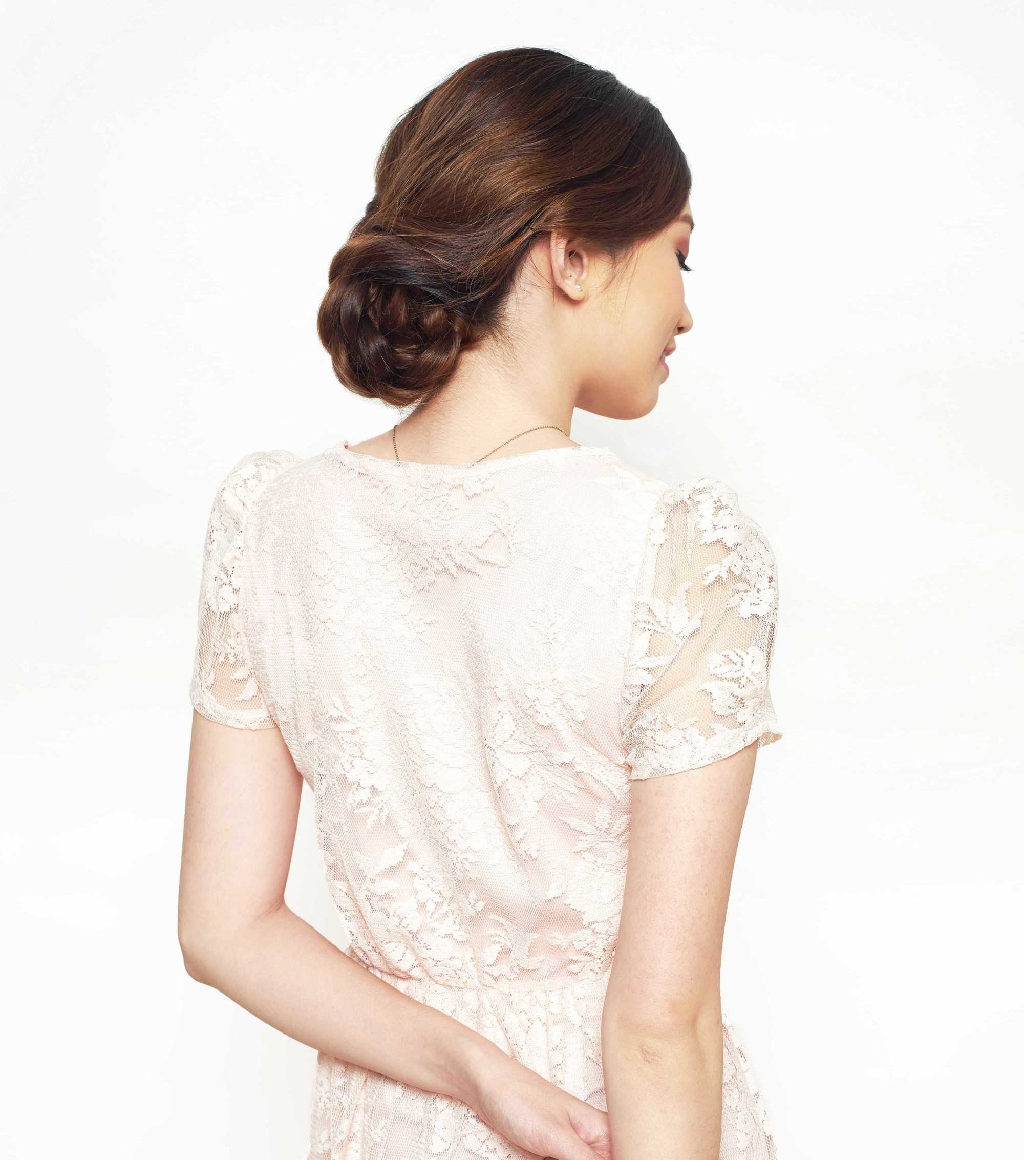 Easy prom hairstyles: Back shot of a model with chestnut brown hair styled into a low braided updo, wearing a white lace dress and posing in a studio.