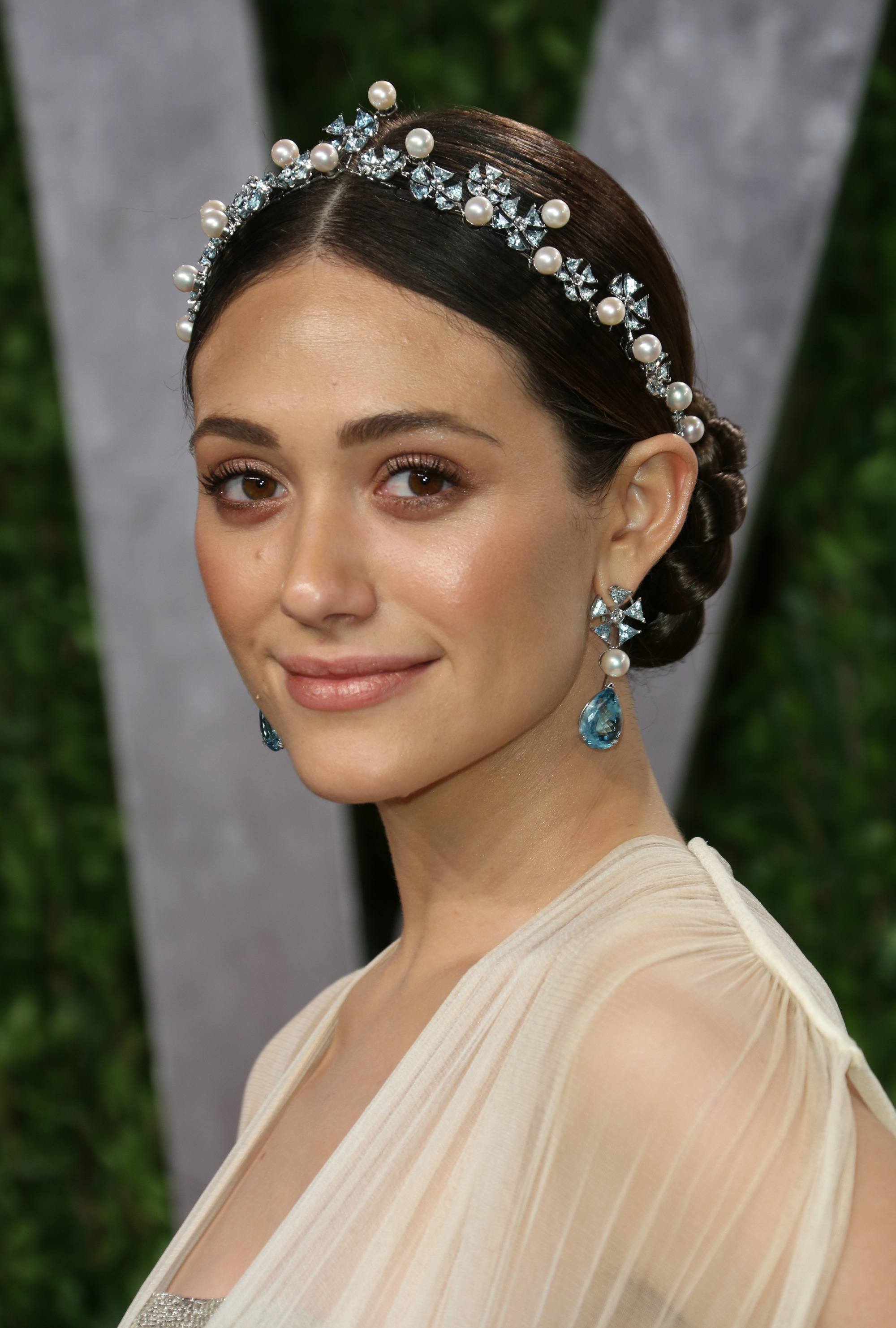 Easy prom hairstyles: Front shot of Emmy Rossum with dark brow hair styled into a low braided bun, complete with a pearl tiara hair accessory, wearing all white and posing on the red carpet.