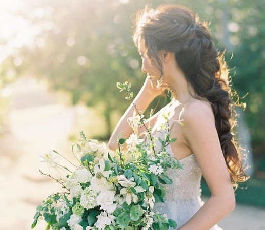 Wedding updos for long hair: Side shot of a bride with long brown hair styled into intricate braided ponytail, wearing a white dress and holding flowers, posing outside.