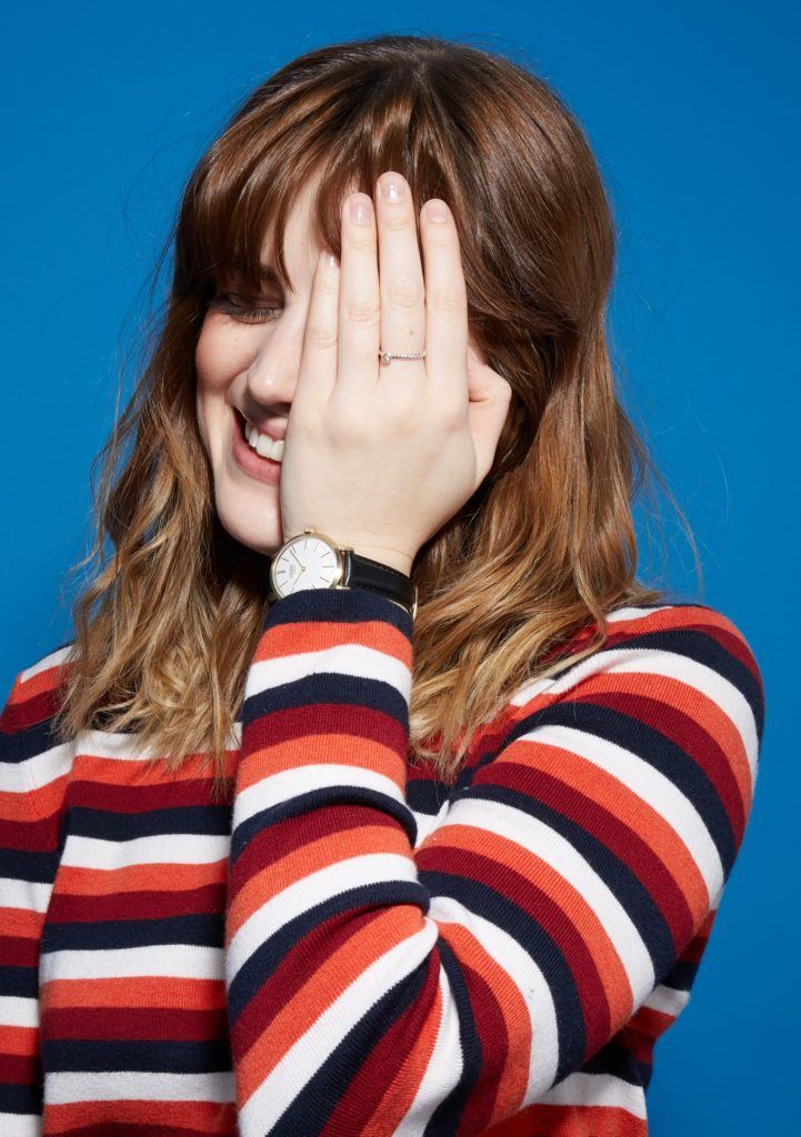woman with medium length wavy brown hair and bangs, touching her hair, touching her face and smiling wearing a striped top