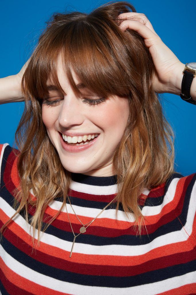 woman with medium length wavy brown hair and bangs, touching her hair and smiling wearing a striped top