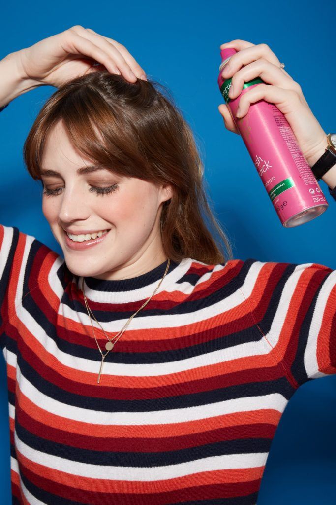 woman with medium length wavy brown hair and bangs, spraying dry shampoo into her hair