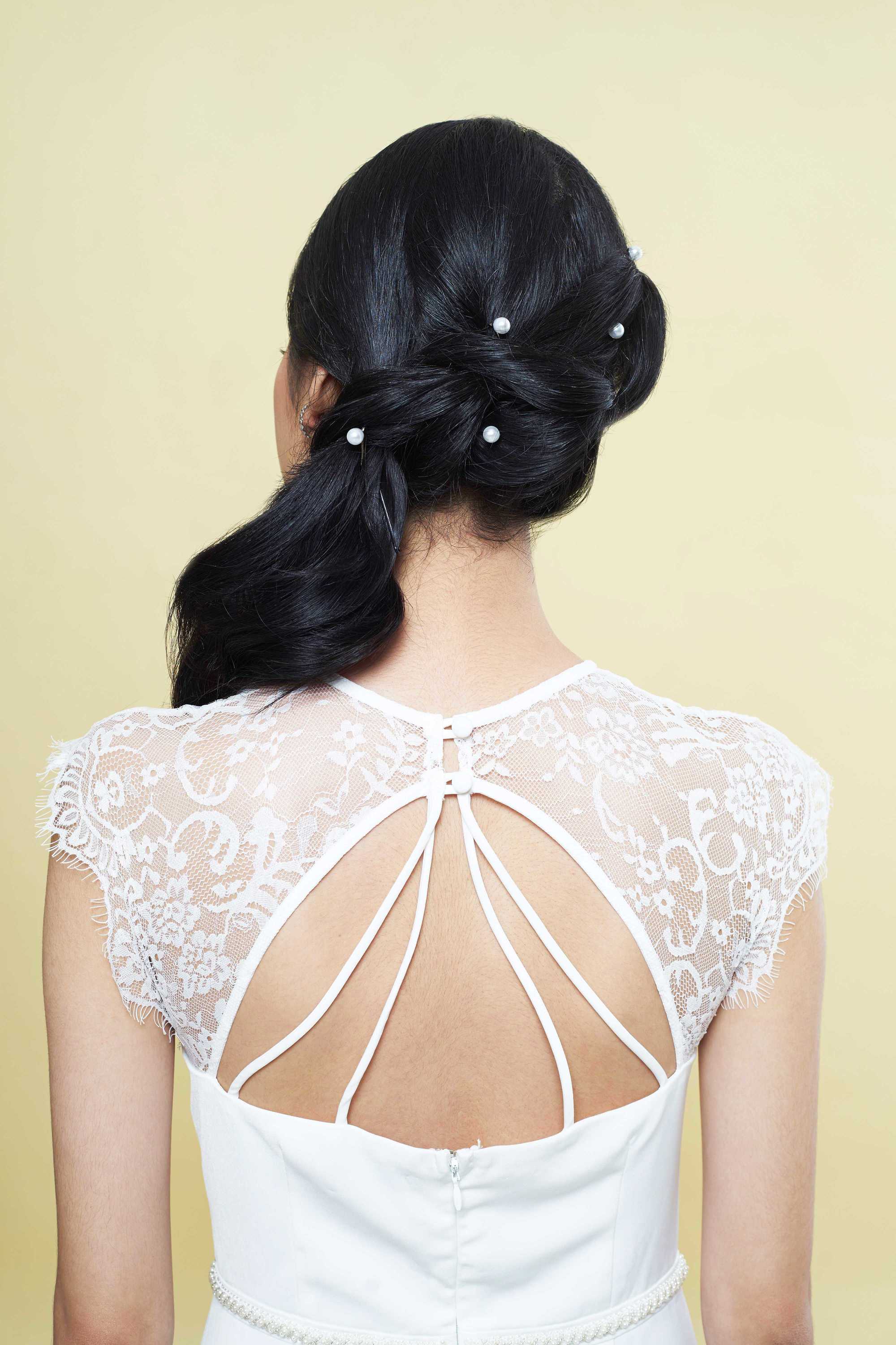Wedding updos for long hair: Back shot of a bride with long dark hair styled into twisted ponytail, complete with pearl hair accessories, wearing a white lace dress and posing in a studio.