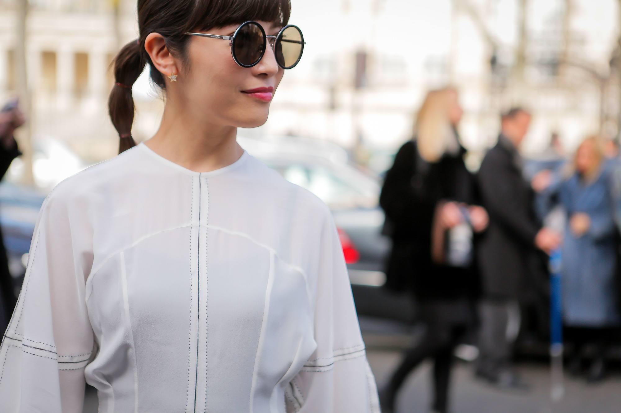 quick updos: street style image of woman with dark brown hair styled in a low bubble ponytail with full blunt bangs wearing a white jacket and round sunglasses