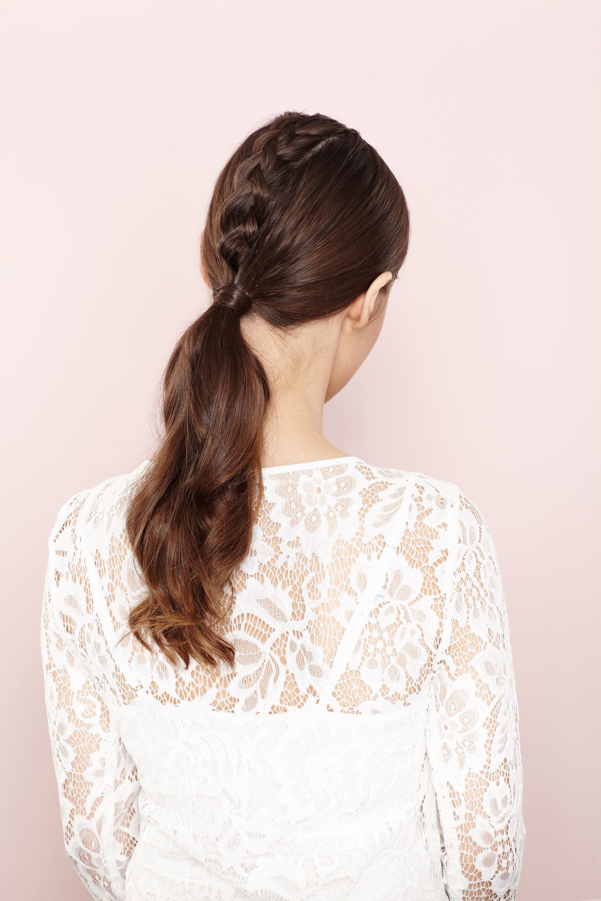 Easy Prom Hairstyles: Back shot of a model with long chestnut brown hair styled into a unicorn braided ponytail, wearing a white lace dress and posing in a studio.