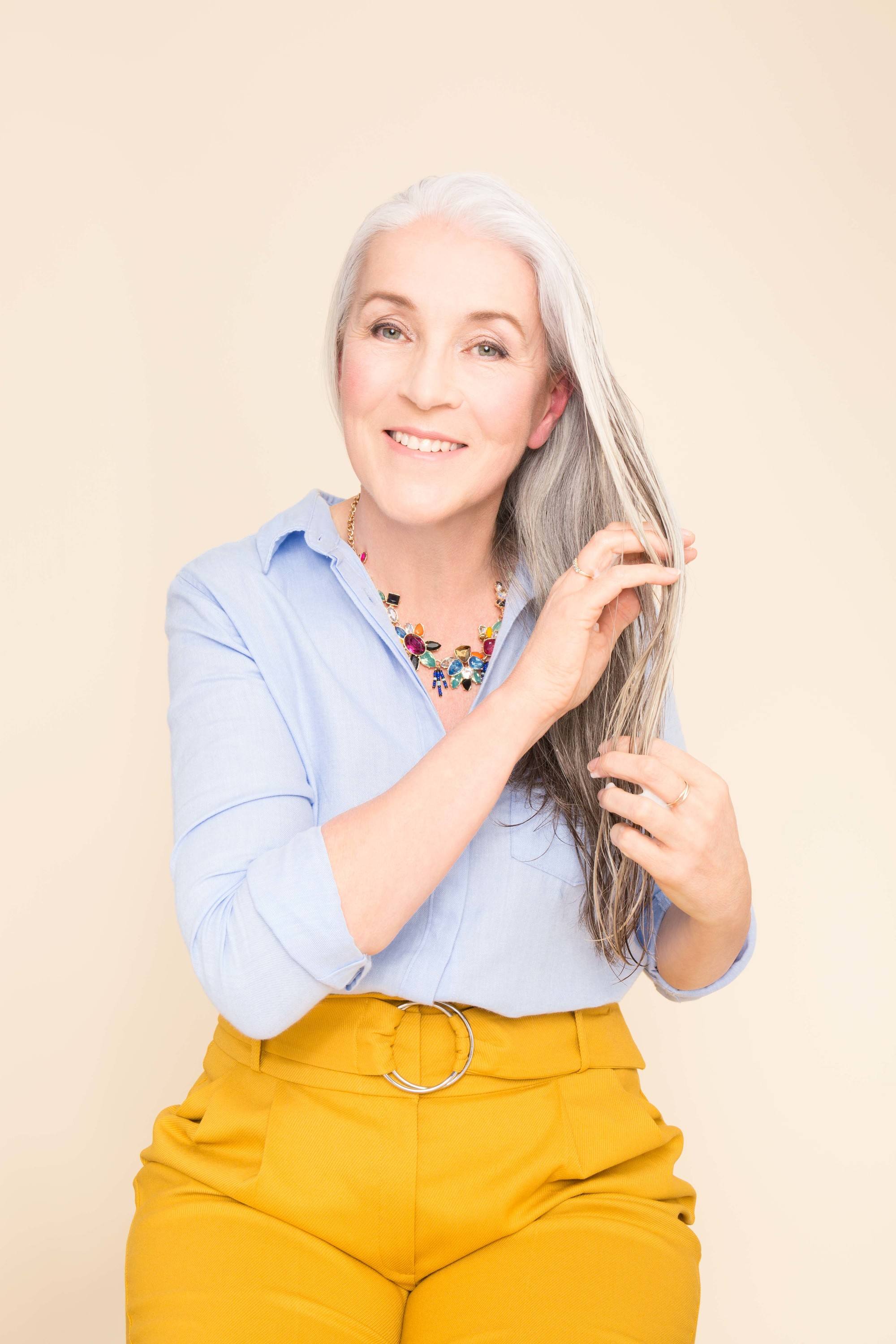 Shampoo full guide: Mature model touching her grey white medium length hair wearing a blue shirt and yellow trousers.