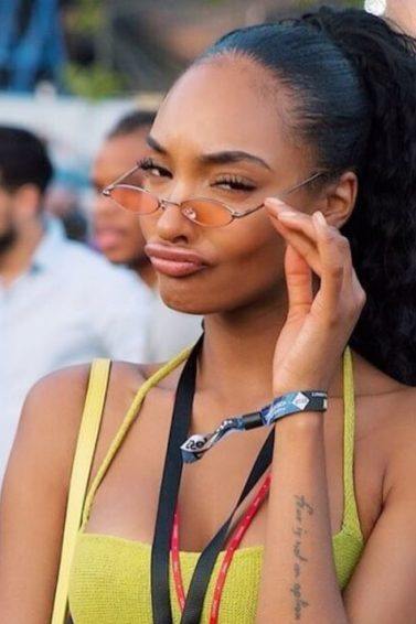 Jourdan Dunn at Wireless Festival 2018 with dark brown hair styled in a high wavy ponytail wearing micro sunglasses and a yellow cami top.