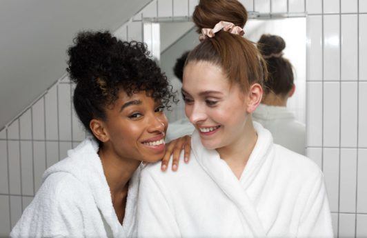 Leave-in conditioner: Shot of two girls in bathrobes, one with natural curly hair in a bun updo, the other with straight hair in a bun, too
