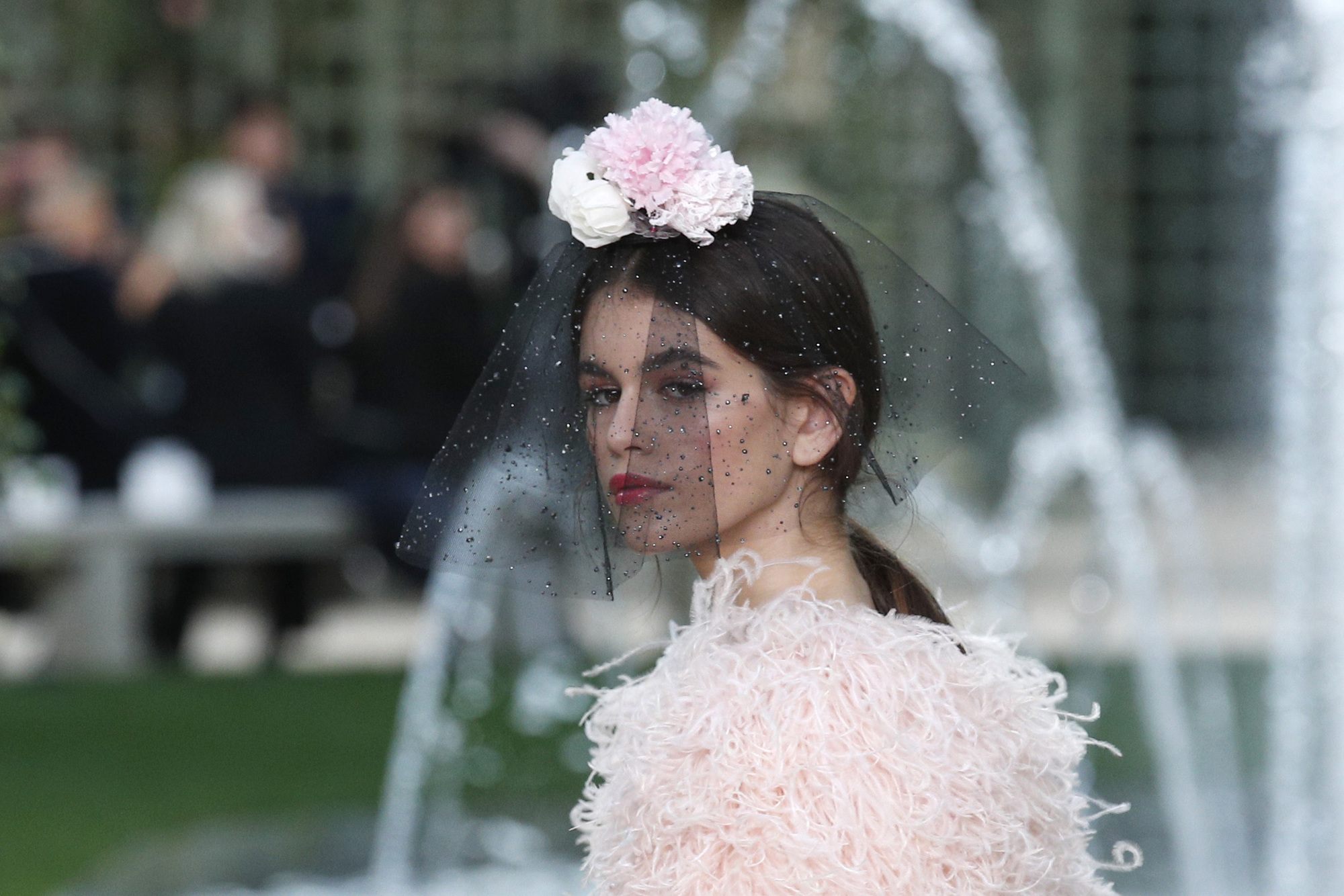 close up shot of kaia gerber with low sleek ponytail with floral viell on it, wearing pink fluffly dress on the chanel paris haute couture runway ss18