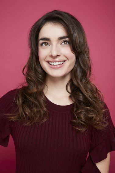 All Things Hair: Woman with romantic long brown curly hair, wearing purple jumper and posing against a pink background
