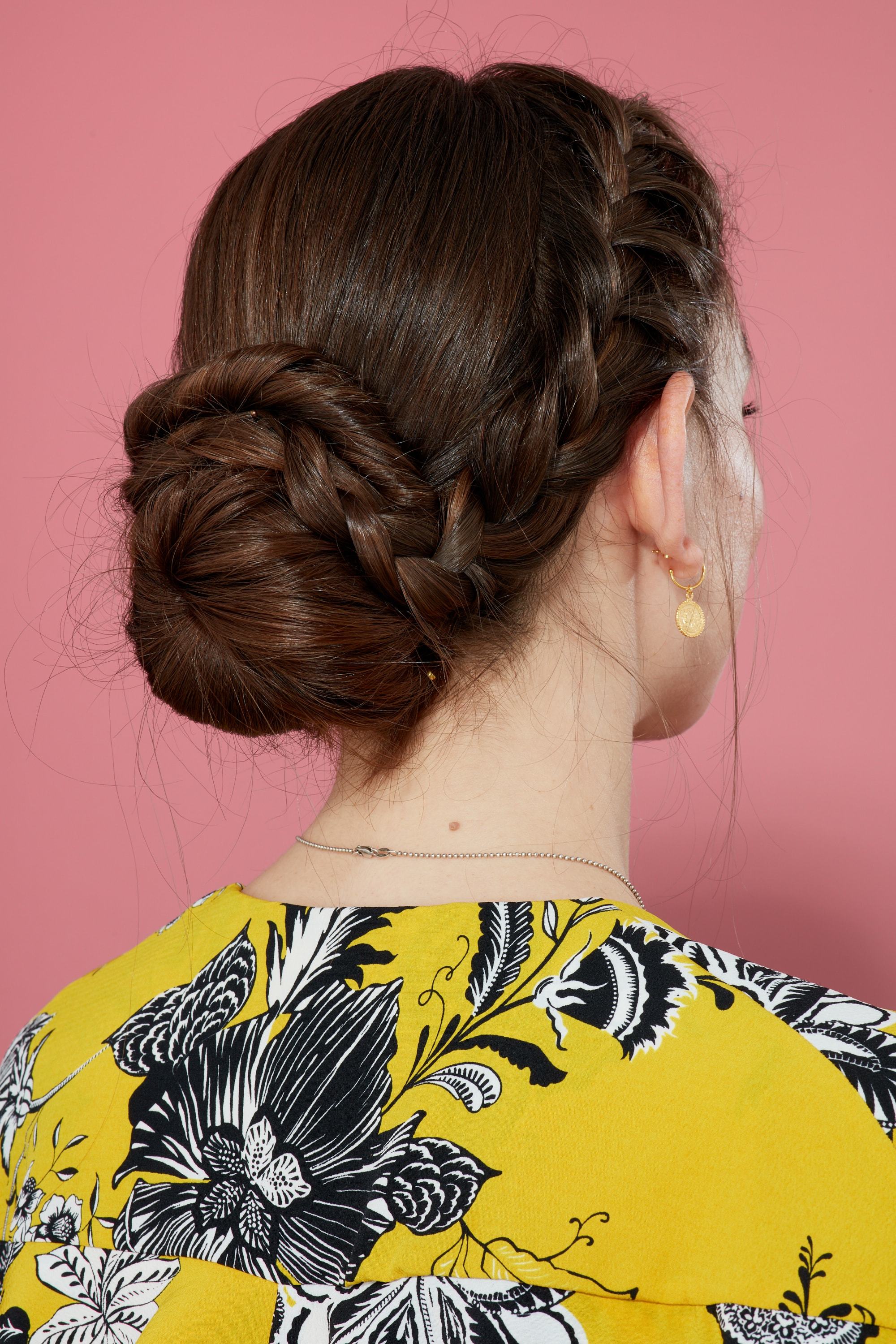 Braids for long hair: Woman with long brown hair in french braided bun wearing yellow pattern top against a pink backdrop.