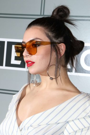 Charli XCX wears her dark tresses in twin bun hairstyle with loose strands falling around her face worn with red sunglasses