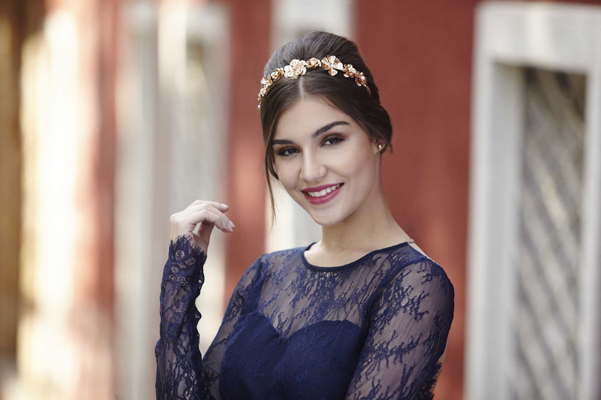 prom hairstyles for medium hair: model with dark brown straight hair styled in a beehive bouffant updo with gold floral headband wearing a navy lace dress