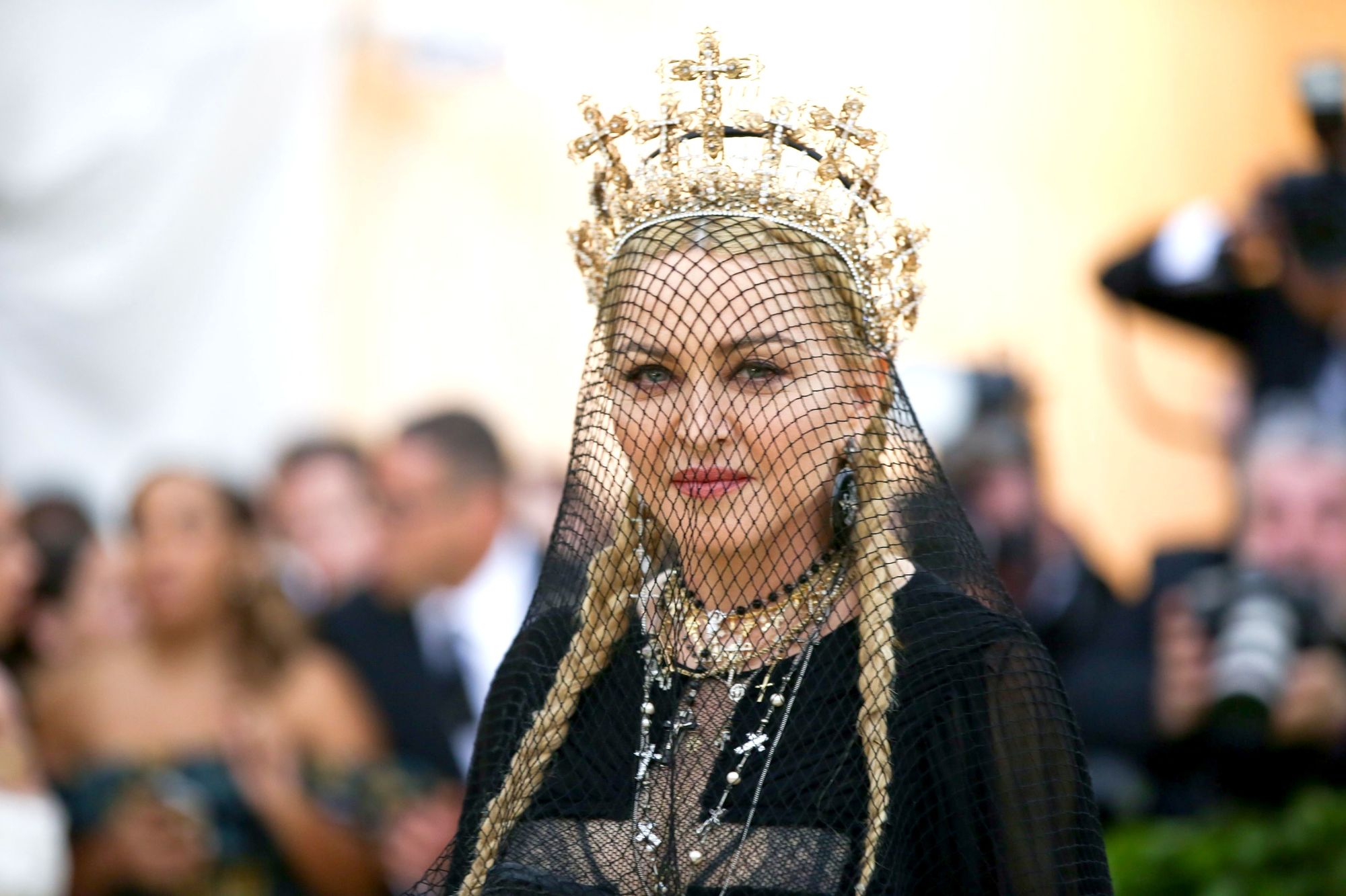 Madonna at the 2018 Met Gala with her blonde hair in two pigtail braids with a black veil and gold crown.