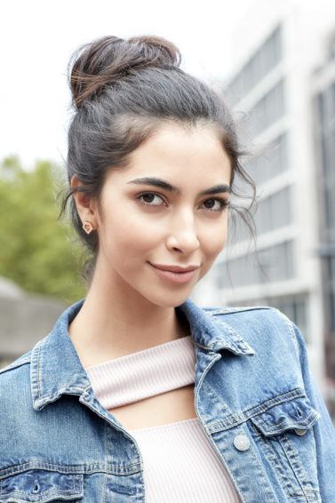 Messy bun tutorial: Brunette woman with her hair in a messy bun, wearing a denim jacket and lilac top, standing outside on the street