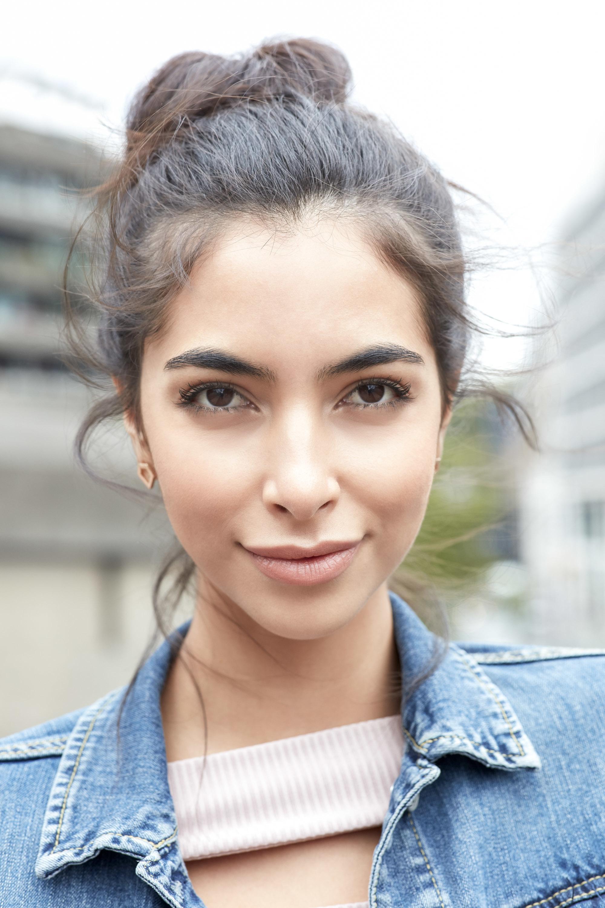 Messy bun tutorial: Close-up outdoor shot of a brunette model with her hair in a messy bun, wearing a denim jacket and lilac top