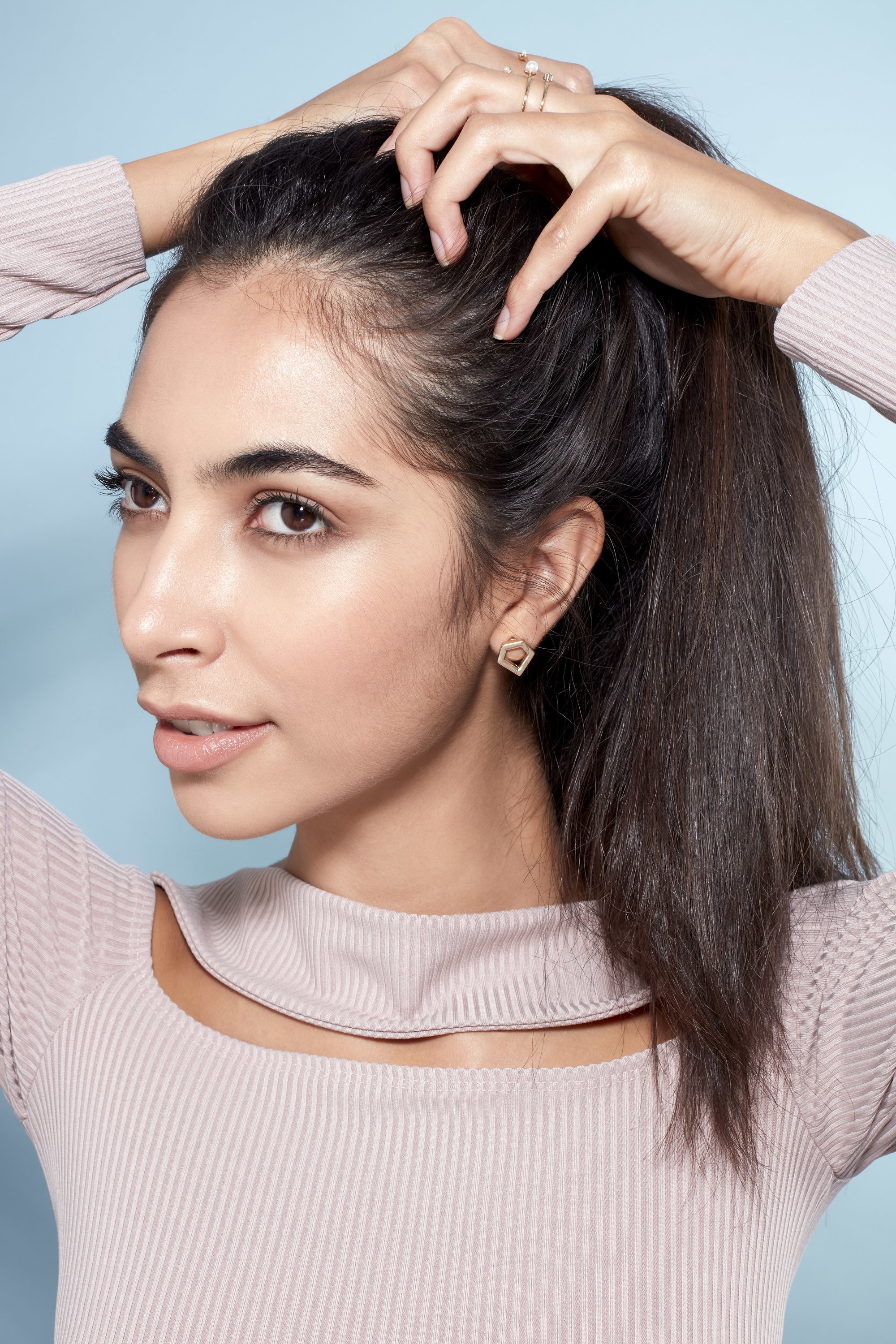 Messy bun tutorial: Close-up of a brunette model with long straight hair raking her hair into a high ponytail, wearing a lilac top and standing against a blue background