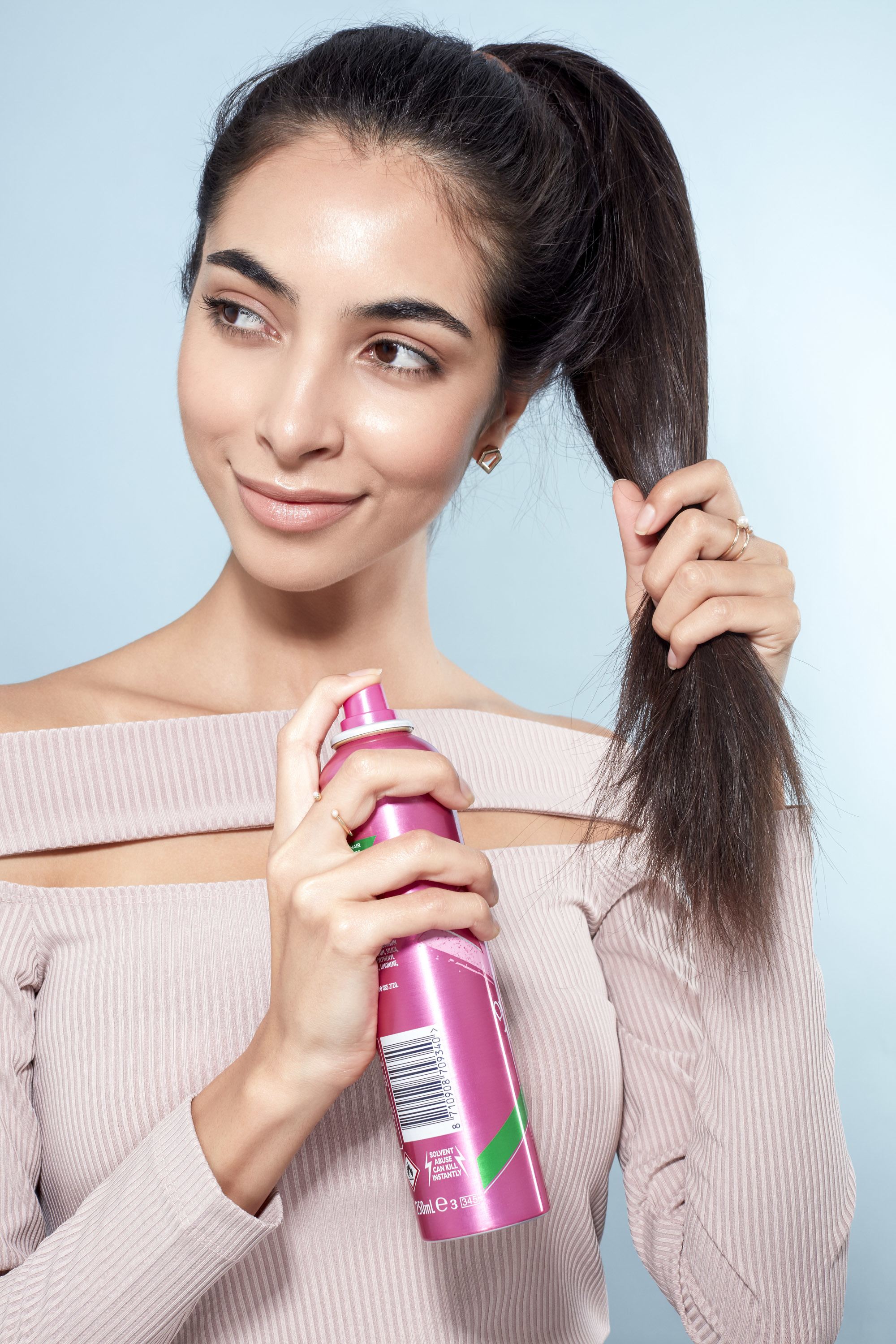 Messy bun tutorial: Close-up of a brunette model with a high ponytail spraying her hair with texturising spray, wearing a lilac top and standing against a blue background