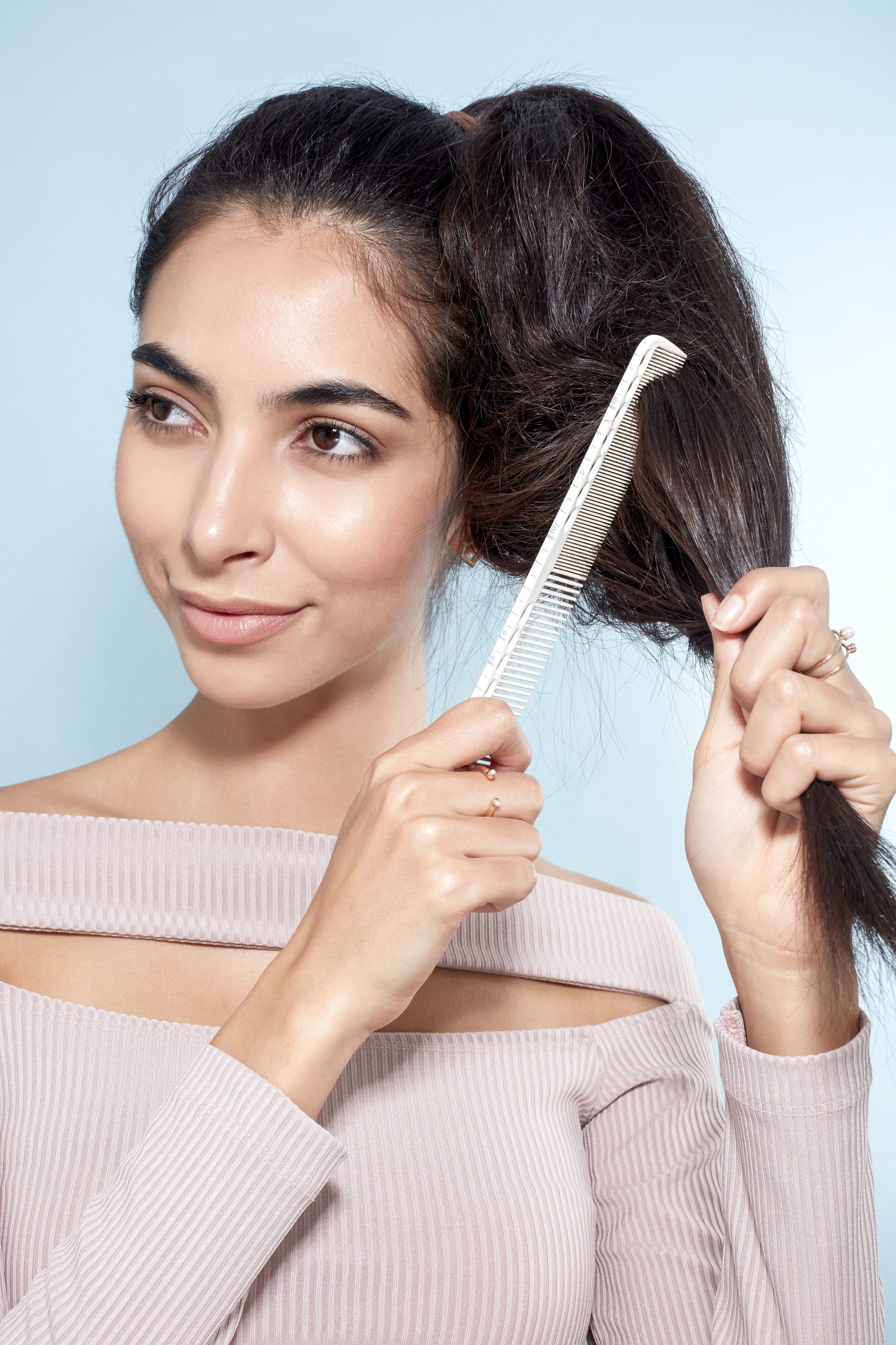 Messy bun tutorial: Close-up of a brunette model backcombing her ponytail with a comb, wearing a lilac top and standing against a blue background