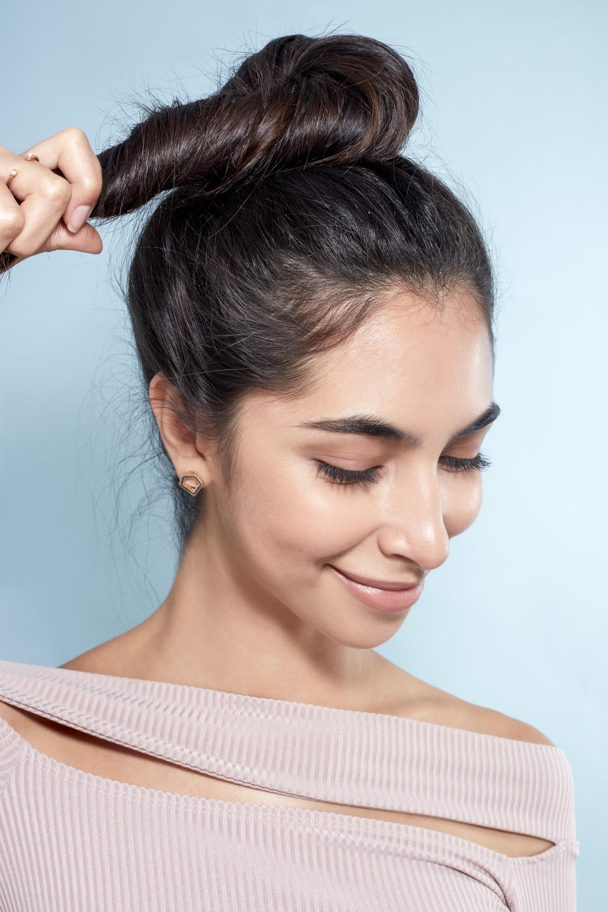 Messy bun tutorial: Close-up of a brunette model wrapping her hair into a high bun, wearing a lilac top and standing against a blue background