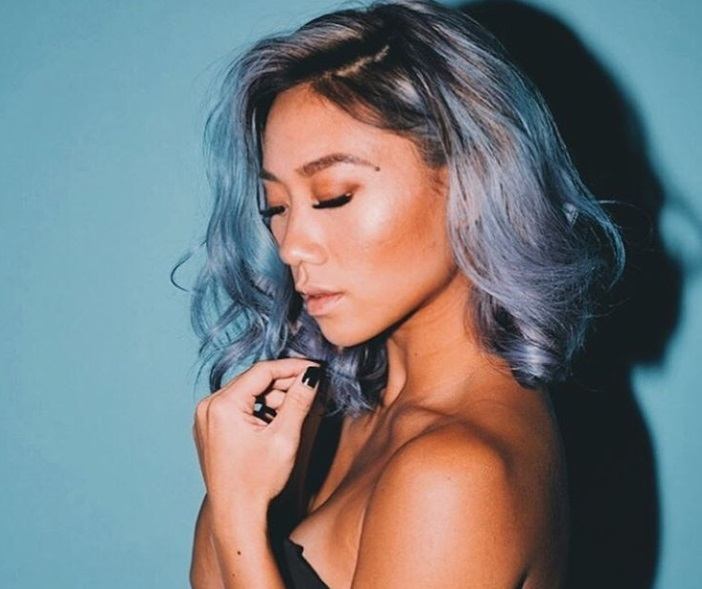 summer hair colour ideas: close up shot of woman with pastel blue wavy hair with dark brown roots, wearing black top and posing against a blue background