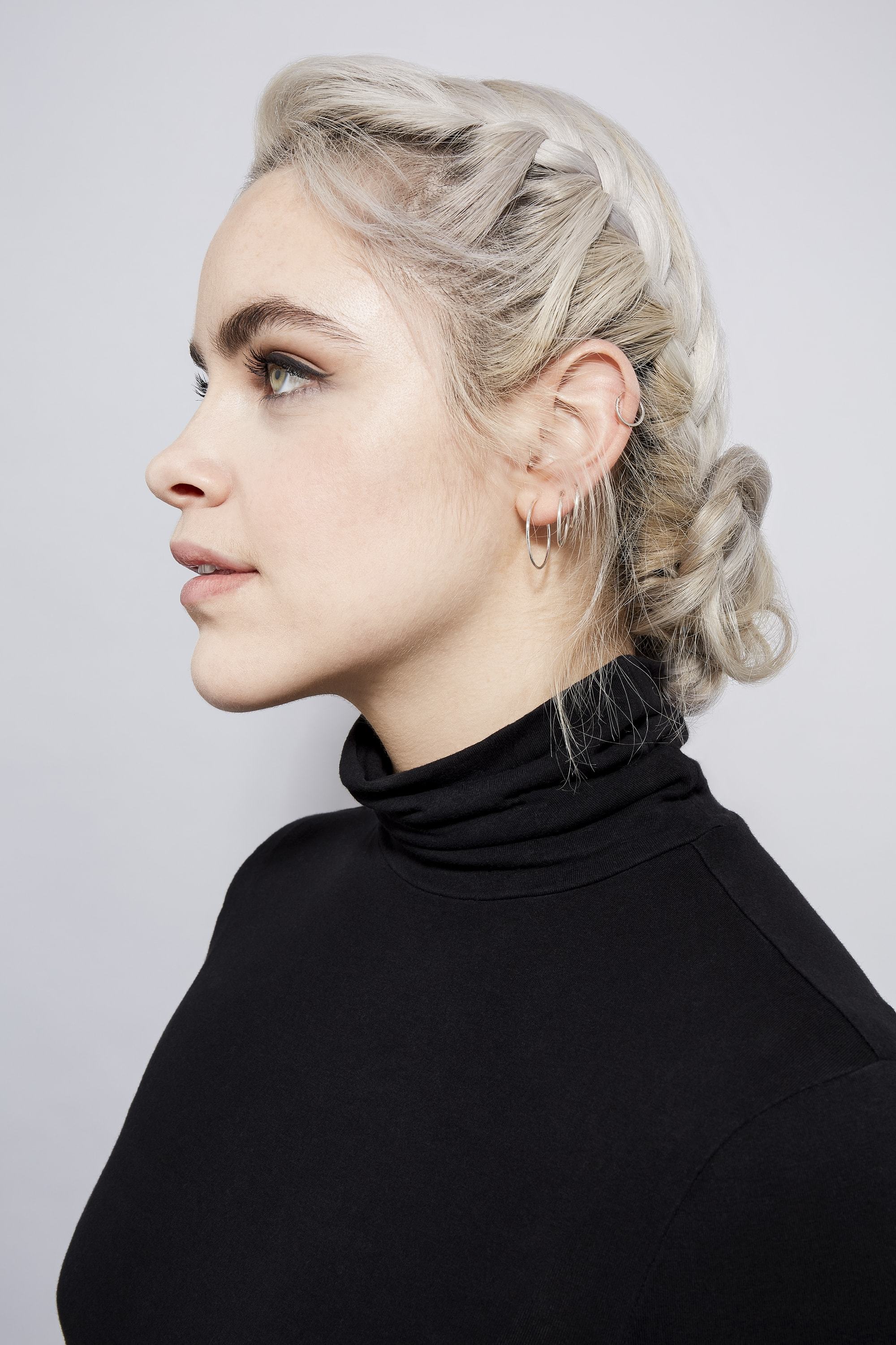 Easy braids for long hair: Side profile of a woman with ash blonde hair in a braided bun, wearing a black roll neck top