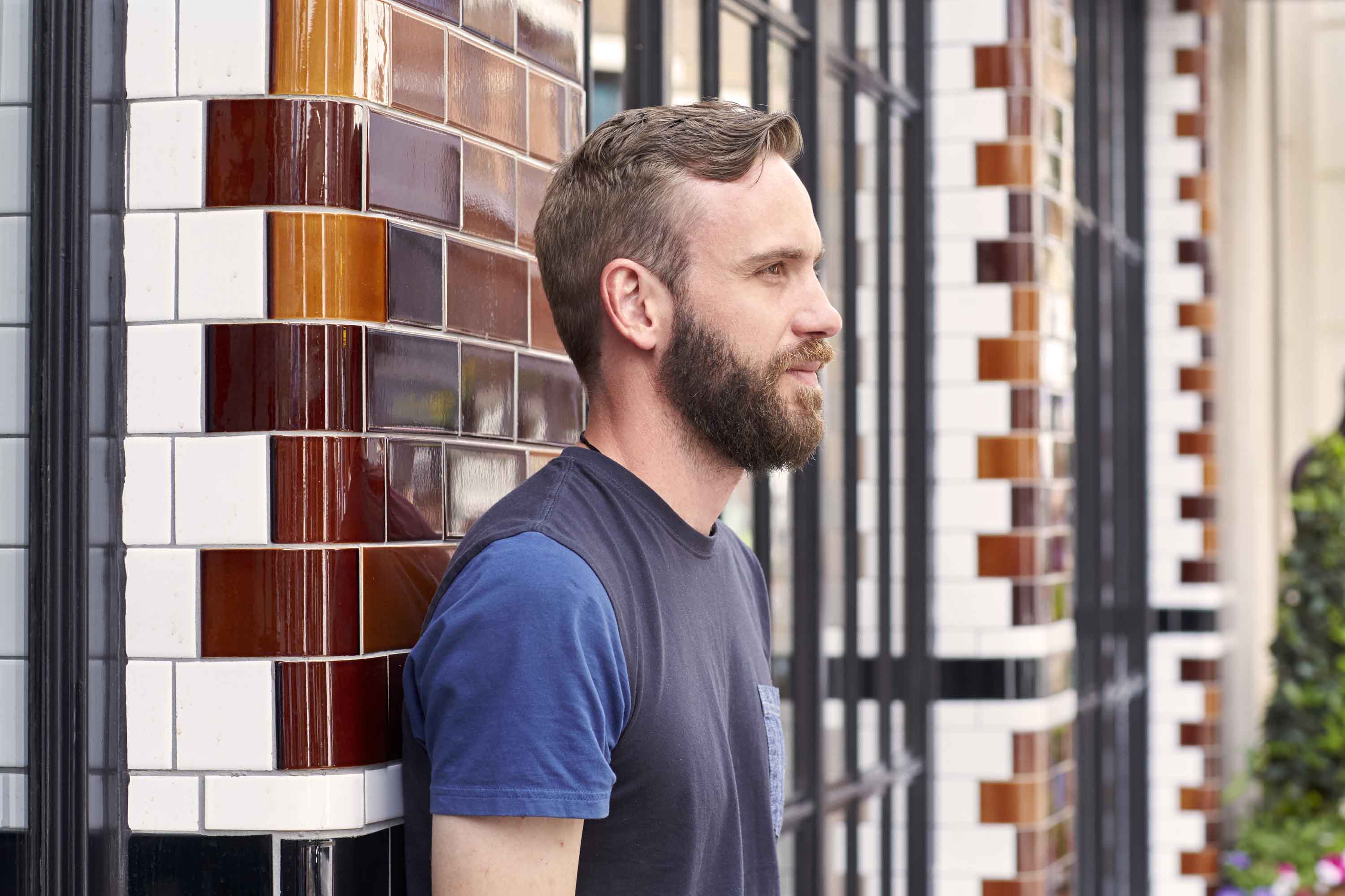brunette man leaning against a wall with a cropped haircut and a beard