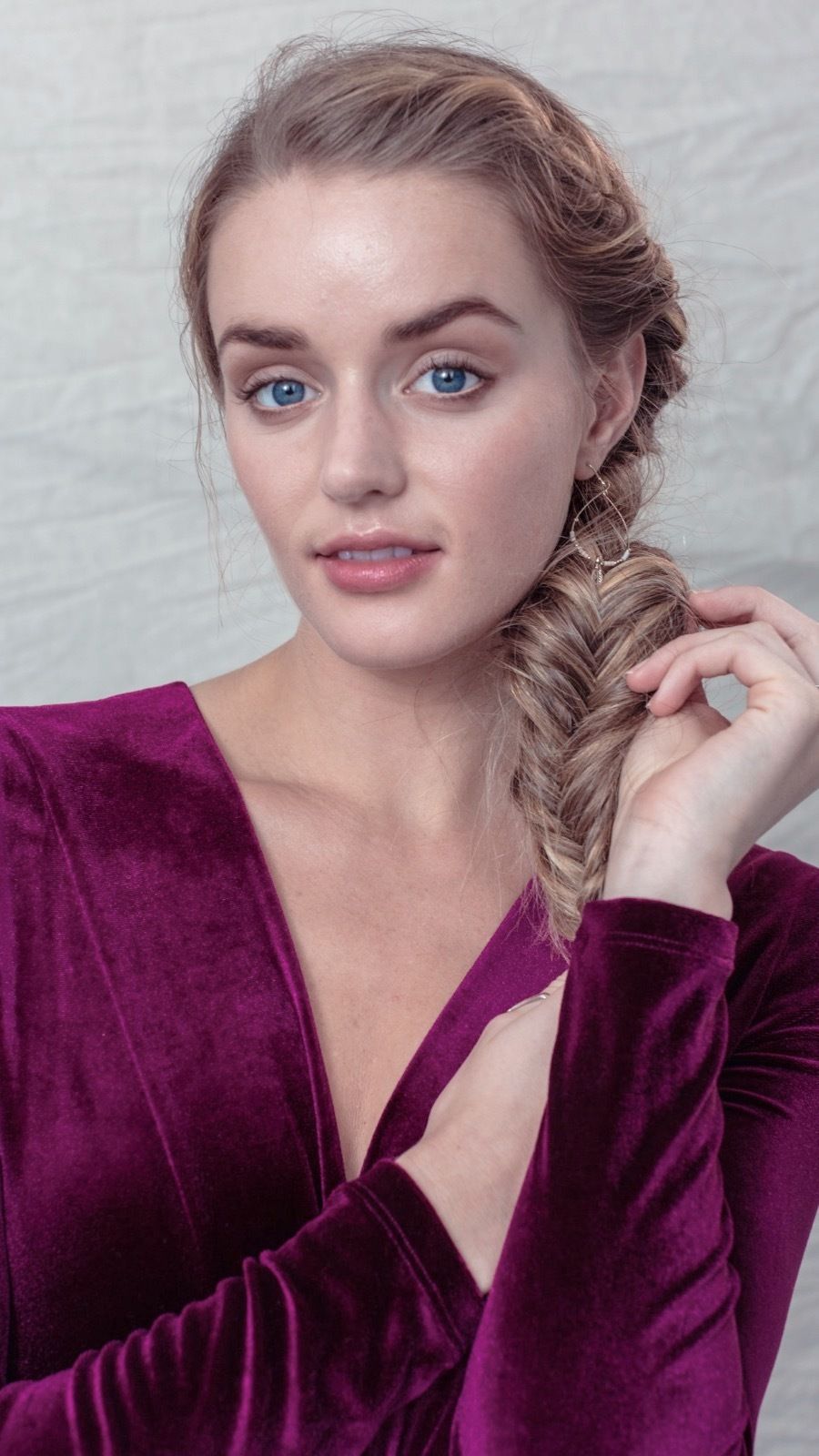 Party hair tips: Woman with long bronde hair styled into a boho fishtail side braid, wearing a purple velvet bodysuit while posing in a studio setting