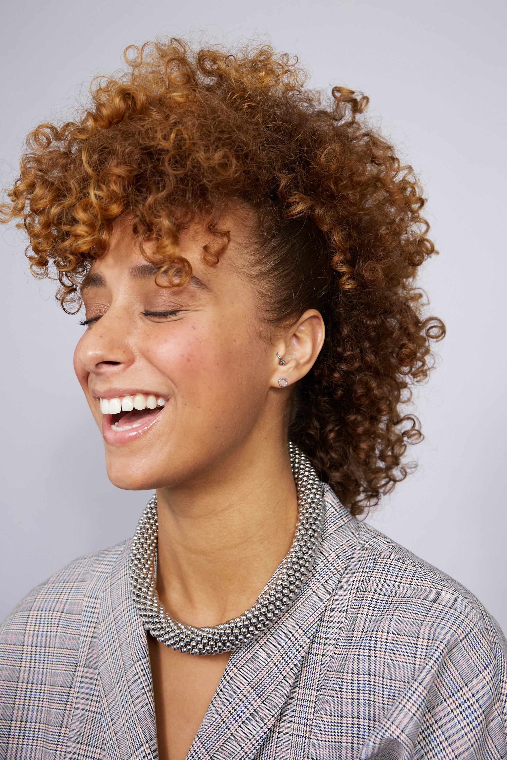 Party hair tips: Woman with natural caramel brown medium afro curls hair styled into frohawk, wearing a checked blazer and a silver necklace