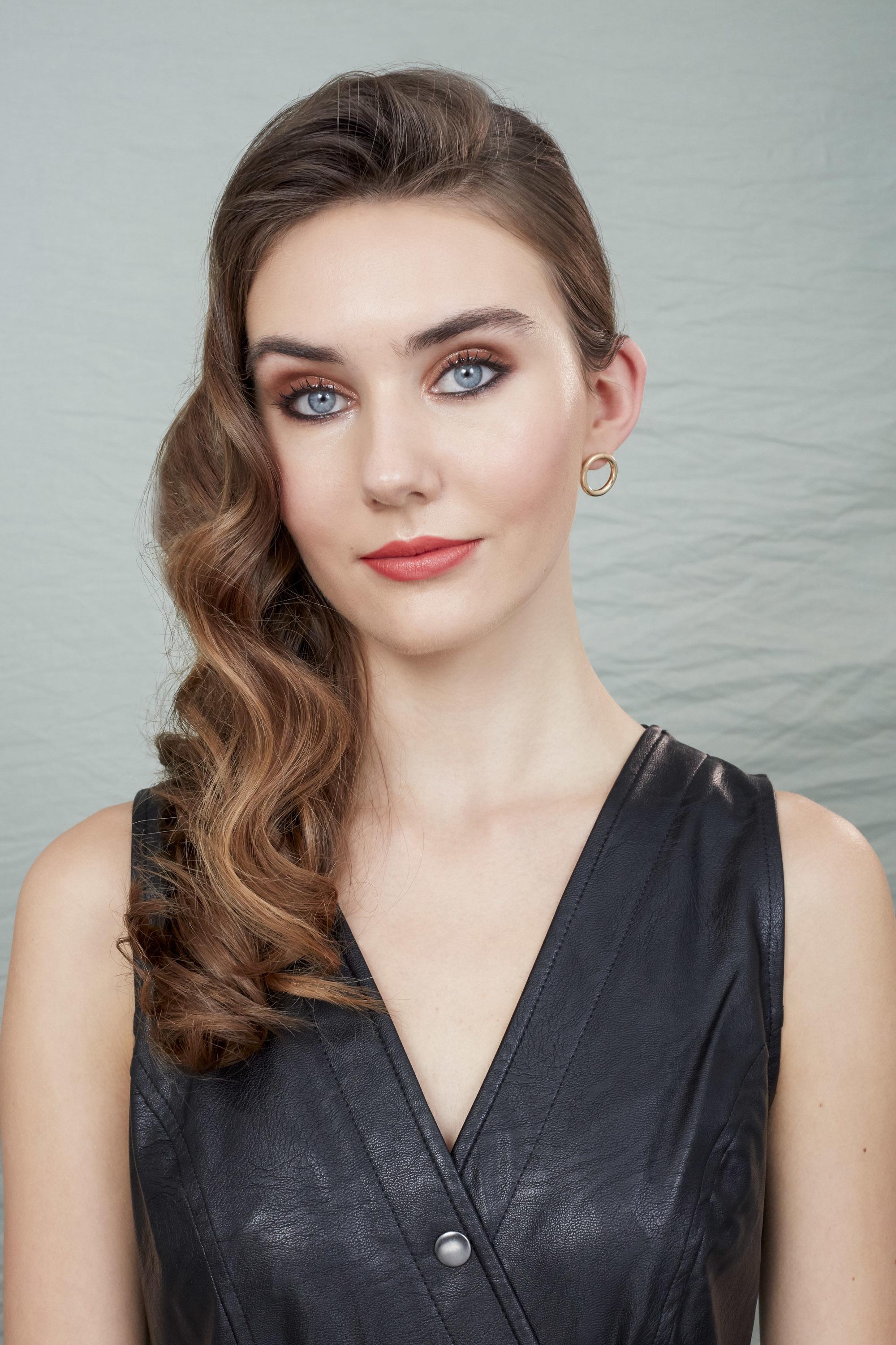 Party hair tips: Woman with long, wavy chestnut brown hair styled into Hollywood curls wearing black leather dress and posing in a studio setting