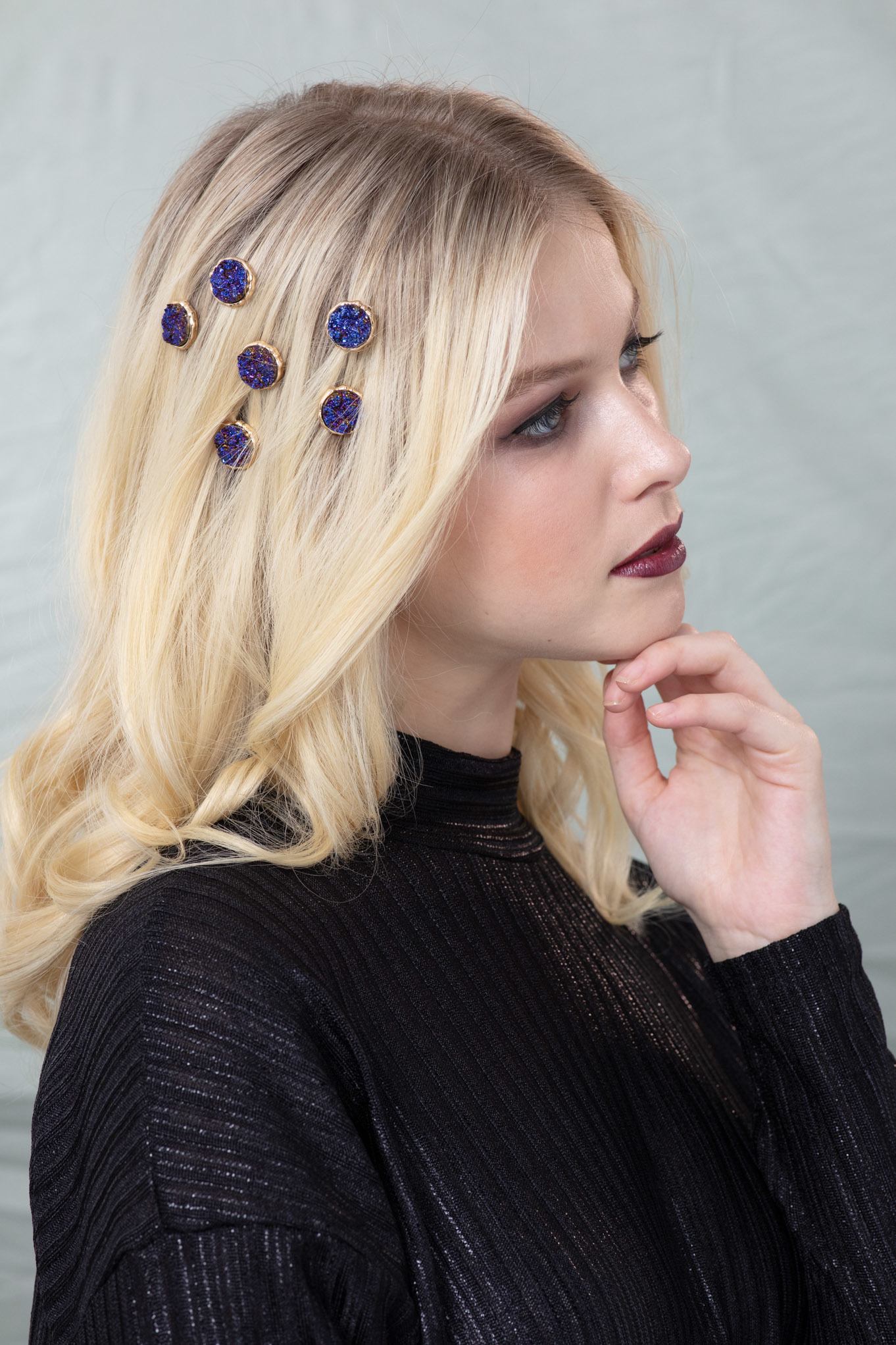 Woman with golden blonde hair styled into loose waves and decorated with Aaron Carlo volcanic hair accessories, wearing a blue top with dark makeup in a studio setting