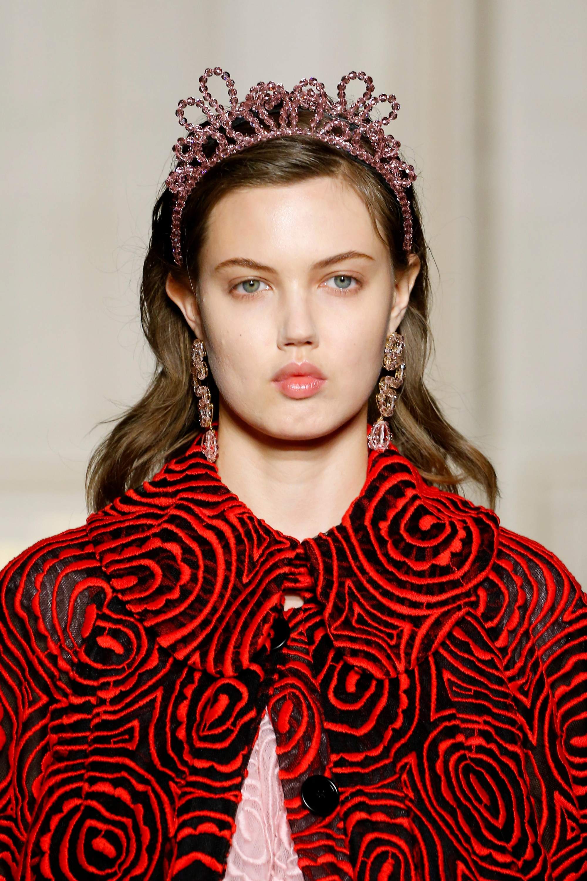 London Fashion Week AW19: Model at Simone Rocha with wavy brunette hair wearing a pink beaded headband in the shape of a tiara, wearing a red coat