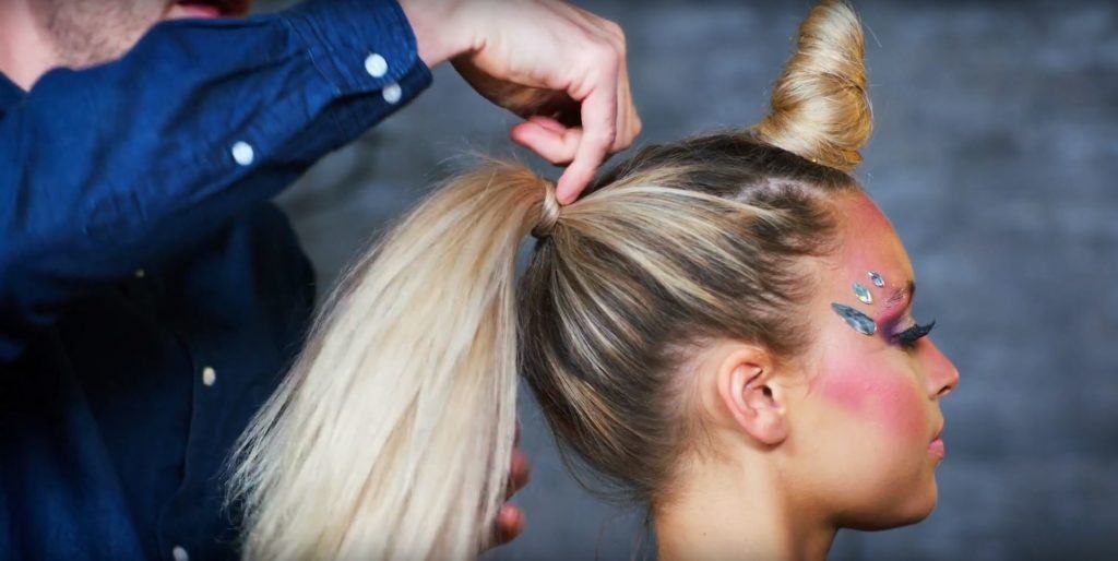 Unicorn Halloween tutorial: Close-up of a hairstylist tying a blonde model's hair in a ponytail, with a unicorn horn inspired bun detail