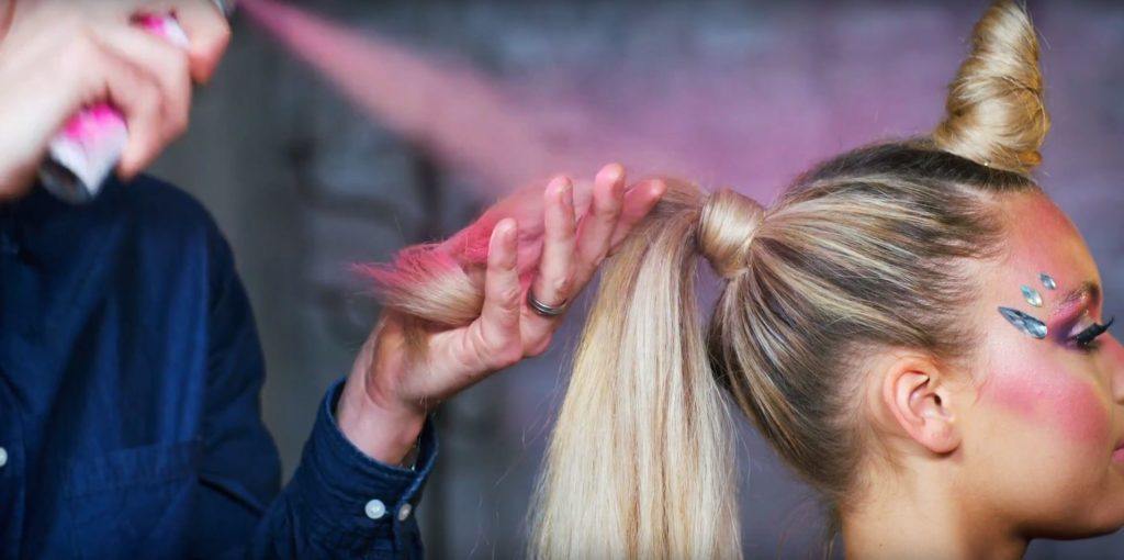 Unicorn Halloween tutorial: Close-up of a blonde model with a unicorn inspired hair tutorial being sprayed with pink coloured hair spray