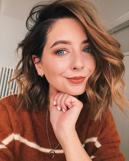 Fall hair colours: Zoe Sugg Zoella with a cinnamon wavy bob wearing a brown jumper resting her chin on her hand.