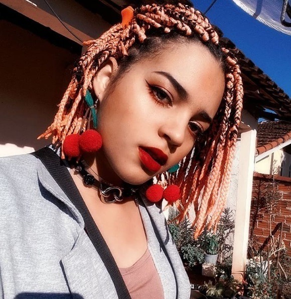 Bob braids bob: Woman with rose gold short box braids, wearing cherry earrings with grey blazer and pink top, posing outside
