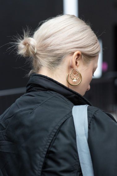 Woman with short bleached blonde hair styled into a low bun, wearing black jacket with hoop earrings on the street