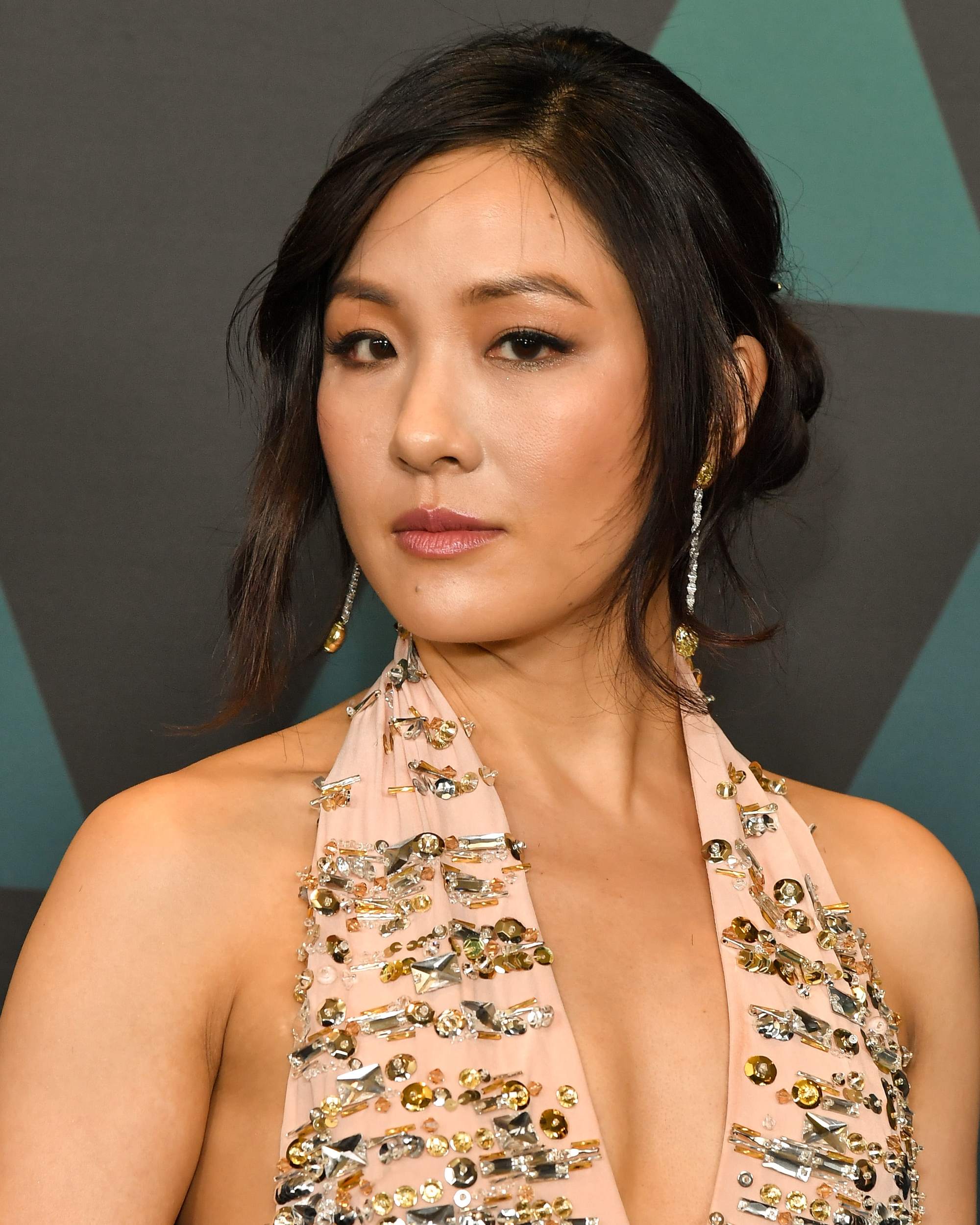Constance Wu with dark brown hair with caramel highlights, styled into a low bun with loose tendrils at the front, wearing a pink and gold halterneck top with drop earrings