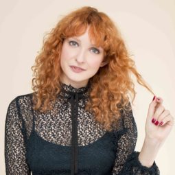 front view of red head model with full curls and fringe with strand of hair in her hand