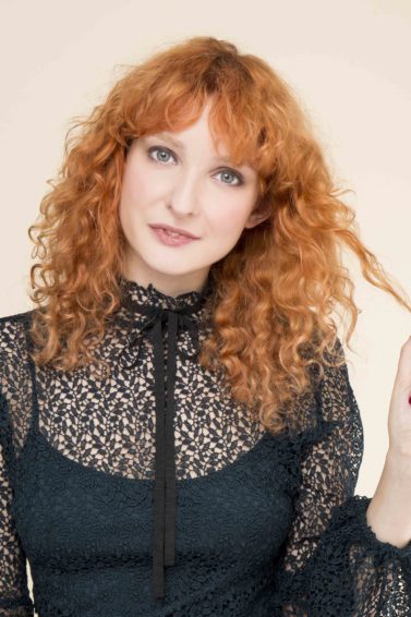 front view of red head model with full curls and fringe with strand of hair in her hand