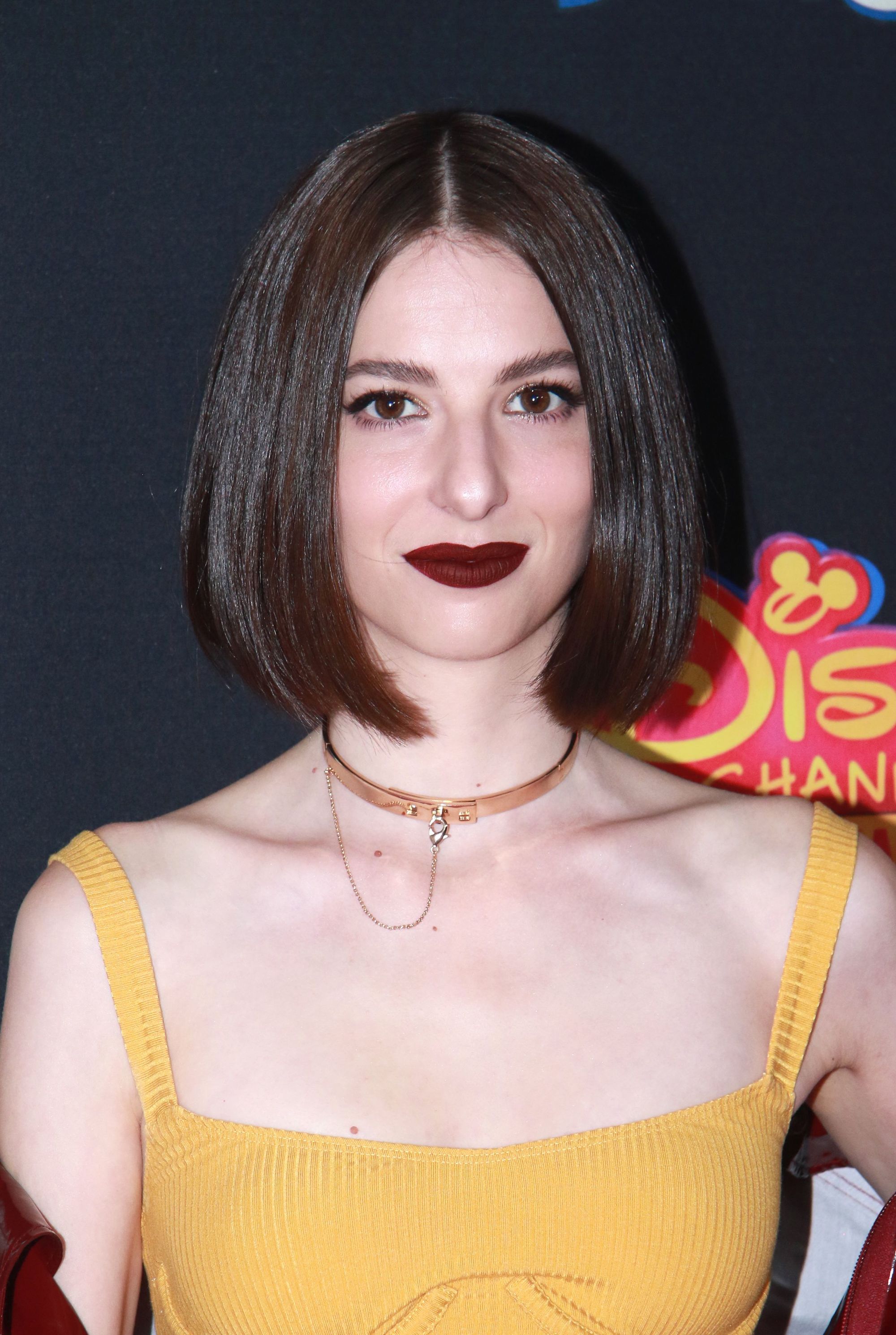 Genevieve Buechner with bob-length brown bronze dark hair styled straight, wearing a yellow top with golden necklace on the red carpet