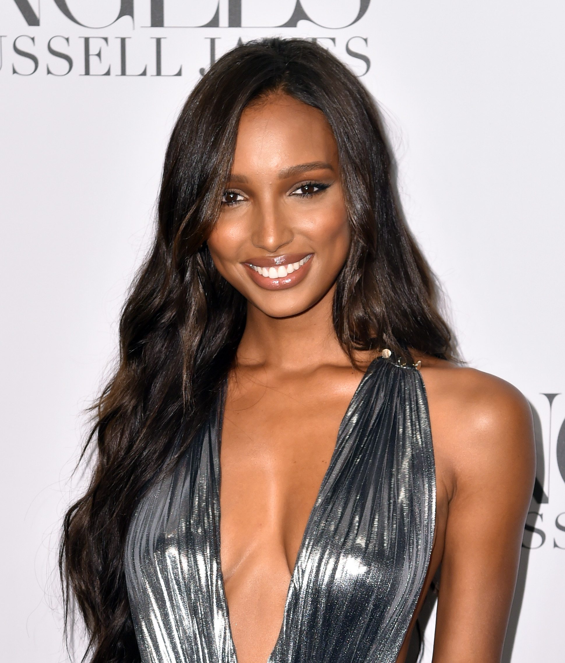 Jasmine Tookes with long, wavy chocolate dark hair with caramel highlights, wearing a silver halterneck dress on the red carpet