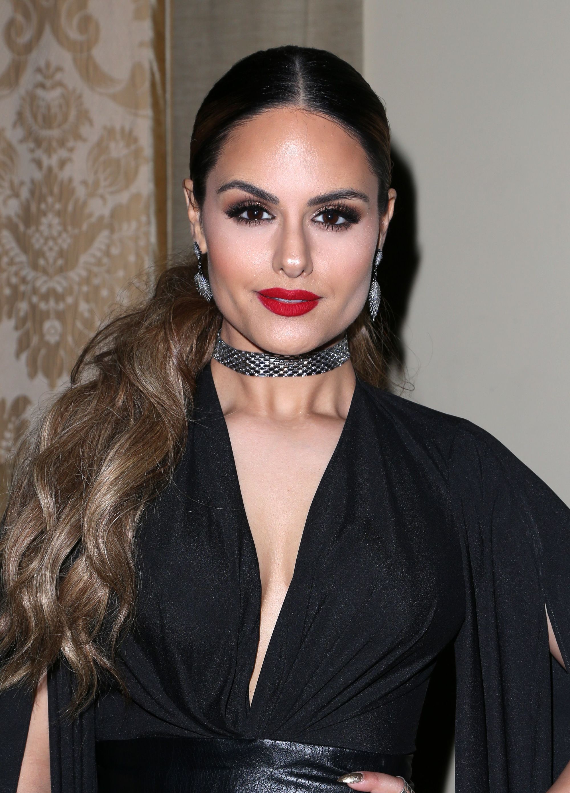 Pia Toscano with long, wavy ash brown hair styled into a ponytail, wearing a black bodysuit with silver choker and earrings on the red carpet