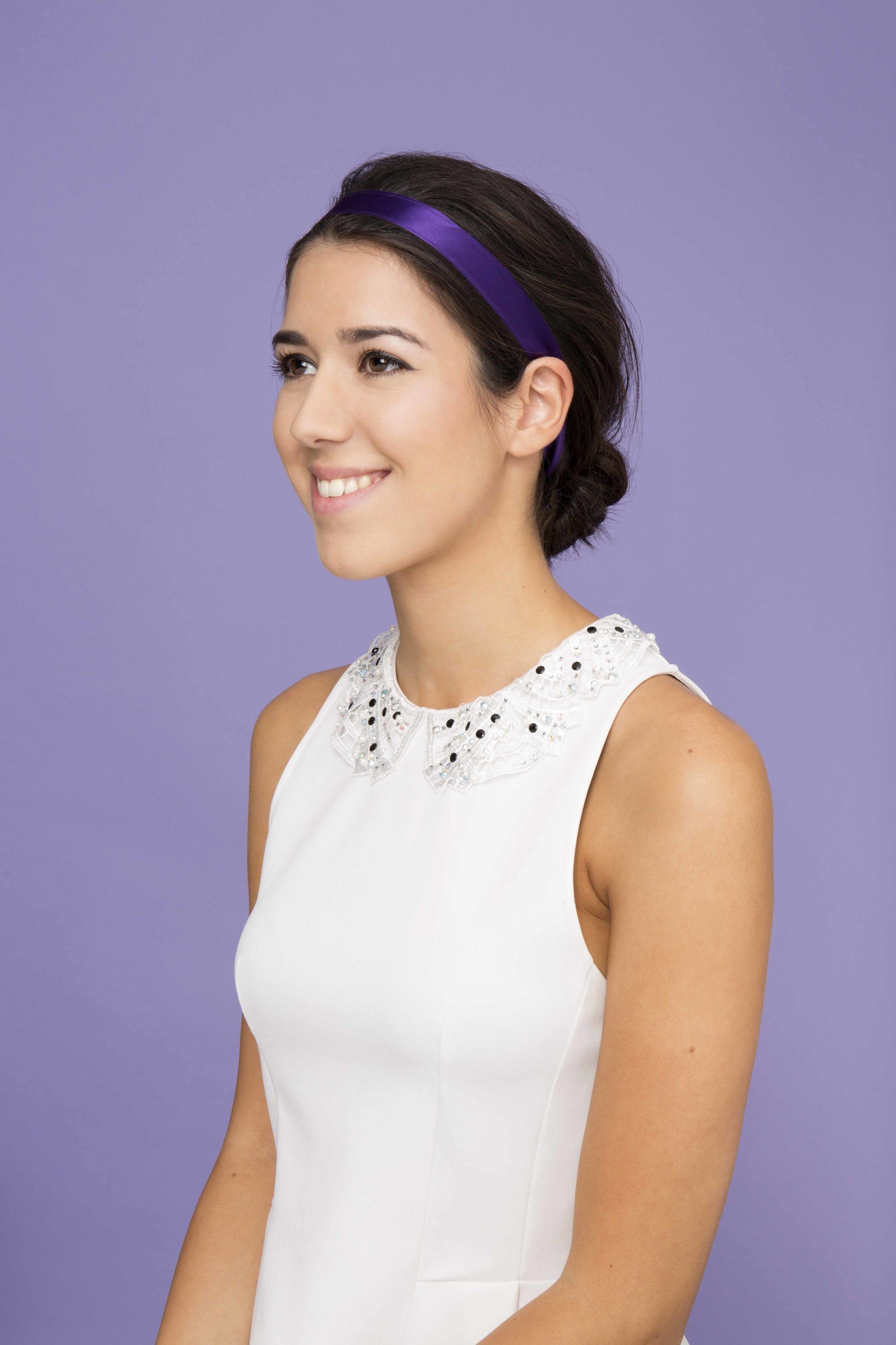 Party hairstyles for medium hair: side Brunette model with hair in mini bouffant and purple ribbon headband wearing a white top against a purple backdrop.