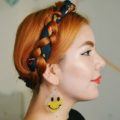 Close up shot of woman with long ginger hair styled into a mikmaid braid updo that has a scarf accessory weaved into it, wearing red lipstick and earrings