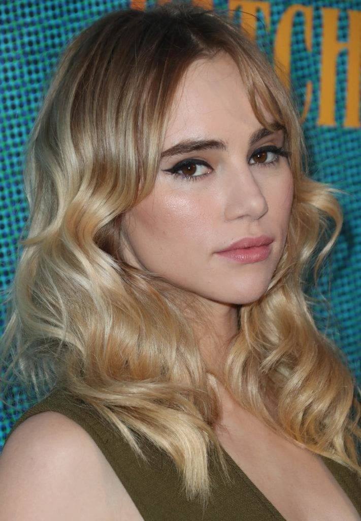 Shades of blonde hair: Suki Waterhouse with wavy blonde balayage medium length hair with long centre parted fringe.